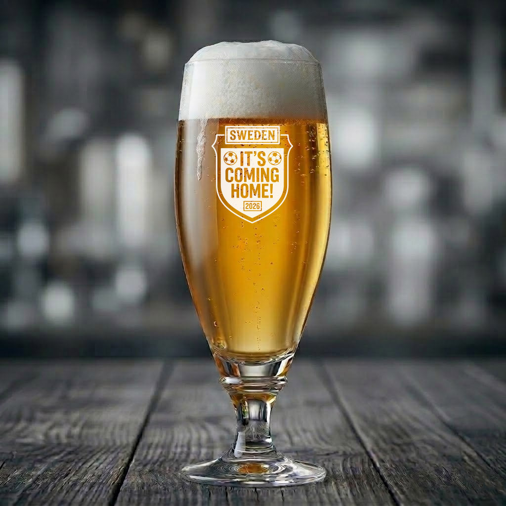 Ett elegant ölglas på fot fyllt med upphälld öl och skumkrona. På glasets framsida finns en vit gravyr formad som en sköld med texten "SWEDEN IT'S COMING HOME!", två fotbollar och årtalet 2026.