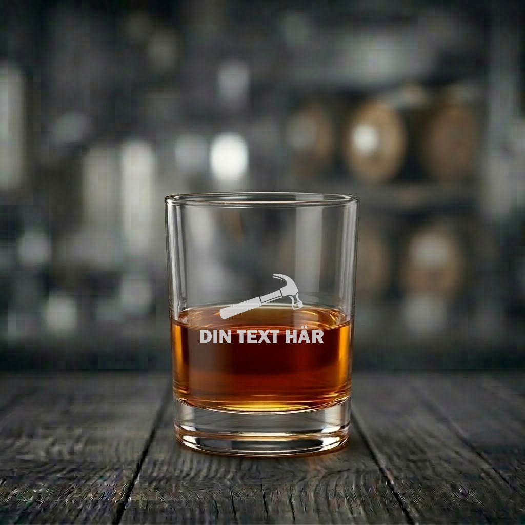 Närbild av ett graverat whiskyglas med ett hammare-motiv och anpassningsbar textfältyta, fyllt med gyllene whisky, stående på en mörk träyta.