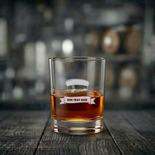 Ett exklusivt graverat whiskyglas fyllt med dryck, med motiv av en klassisk svensk studentmössa och en vimpel med plats för egen personlig text.