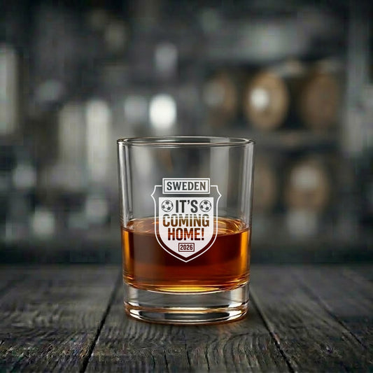 Ett graverat whiskyglas med fotbollsmotiv i form av en sköld och texten "SWEDEN IT'S COMING HOME! 2026", fyllt med whisky och stående på ett rustikt träbord.