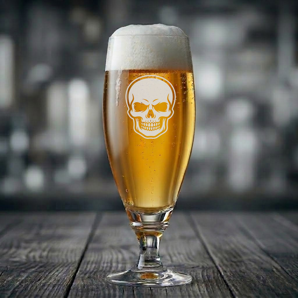 På ett mörkt, väderbitet träbord står ett elegant tulpanformat ölglas fyllt med en bärnstensfärgad dryck och en fluffig, vit skumtopp. Mitt på glaset lyser en krispigt vit lasergravyr av ett detaljerat mänskligt kranium, komplett med markerade tänder och djupa ögonhålor. Miljön bakom glaset är behagligt suddig i dova, industriella nyanser.