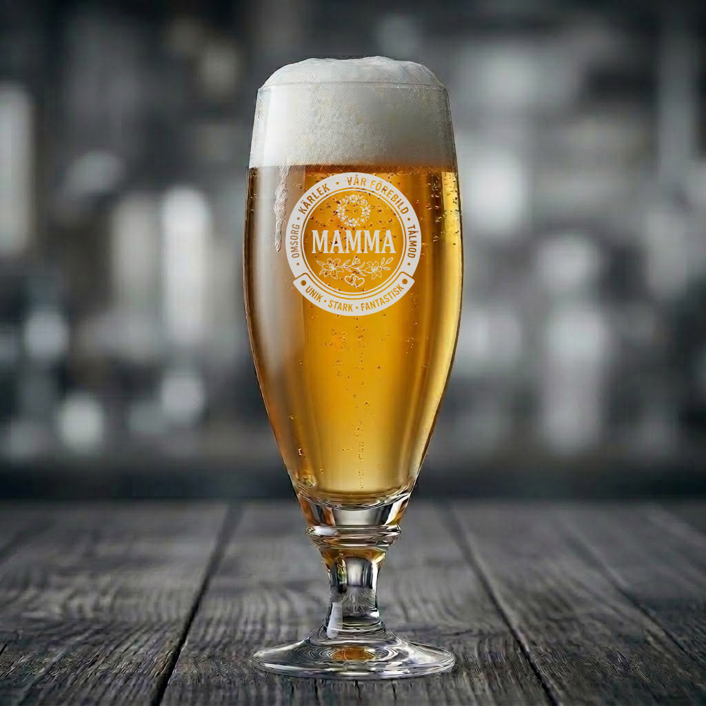 Ett elegant ölglas på fot fyllt med upphälld öl och en skumkrona. Glaset har en vit, cirkelformad gravyr med texten "MAMMA" i mitten inramat av blommor, omgiven av fina ord som "Omsorg", "Kärlek", "Stark" och "Fantastisk".