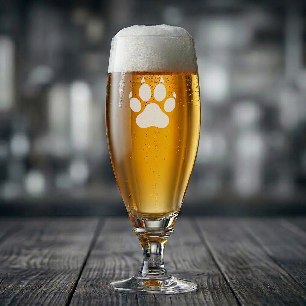 Ett elegant ölglas på fot fyllt med gyllene öl och skumkrona. På glasets framsida finns en stilren vit gravyr som föreställer ett tassavtryck med klor