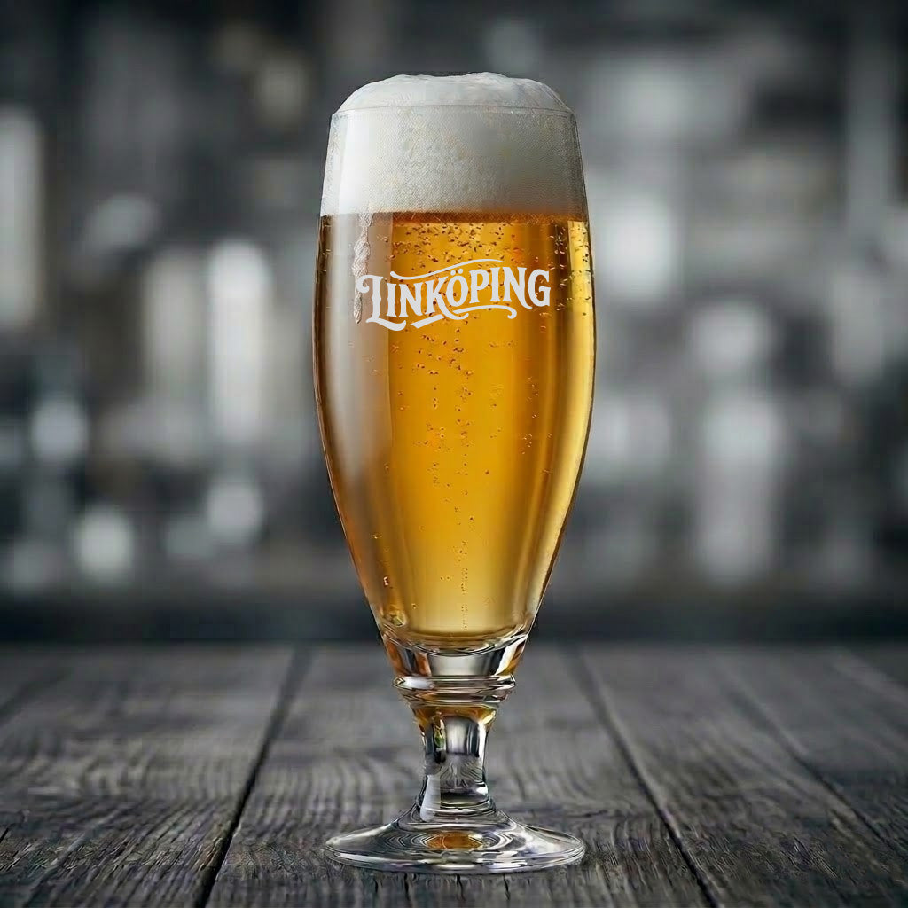 Elegant ölglas med texten Linköping ingraverad, fyllt med gyllene öl med skumkrona på ett träbord.