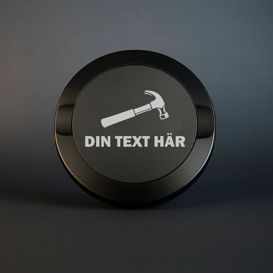 Svart graverad snusdosa i metall med motiv av en hammare och plats för personlig textgravyr.