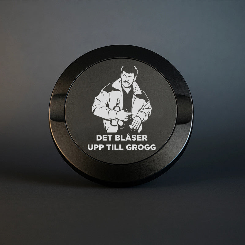 En mattsvart snusdosa i metall med en detaljerad lasergravyr av Christer Pettersson med flaskor och texten DET BLÅSER UPP TILL GROGG, fotograferad mot en mörk och stilren bakgrund.