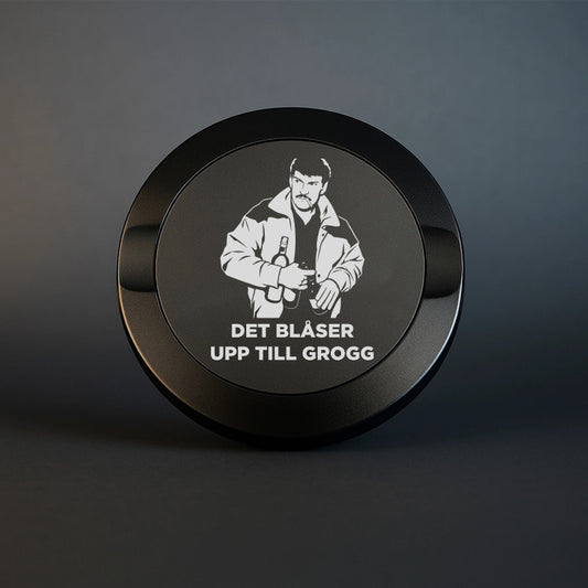 En mattsvart snusdosa i metall med en detaljerad lasergravyr av Christer Pettersson med flaskor och texten DET BLÅSER UPP TILL GROGG, fotograferad mot en mörk och stilren bakgrund.