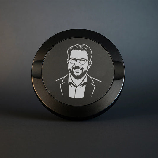 Mattsvart metall snusdosa med detaljerad gravyr av Jimmie Åkesson i kostym och glasögon. Premium snusdosa med porträtt på mörk bakgrund.