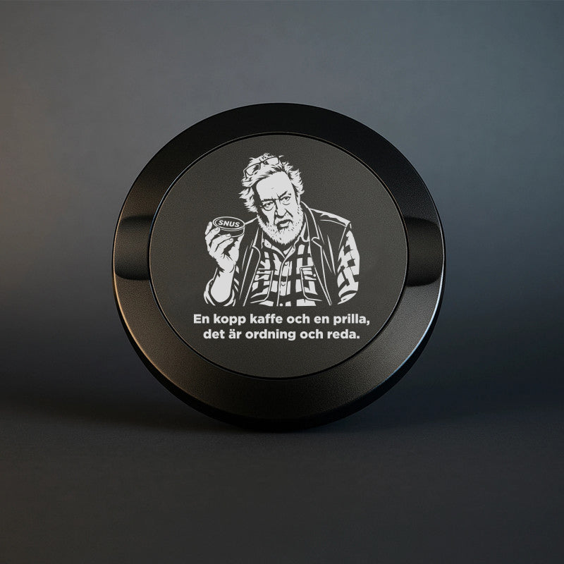 Mattsvart snusdosa i metall med en detaljerad lasergravyr av Leif GW som håller en snusdosa, tillsammans med texten "En kopp kaffe och en prilla, det är ordning och reda".