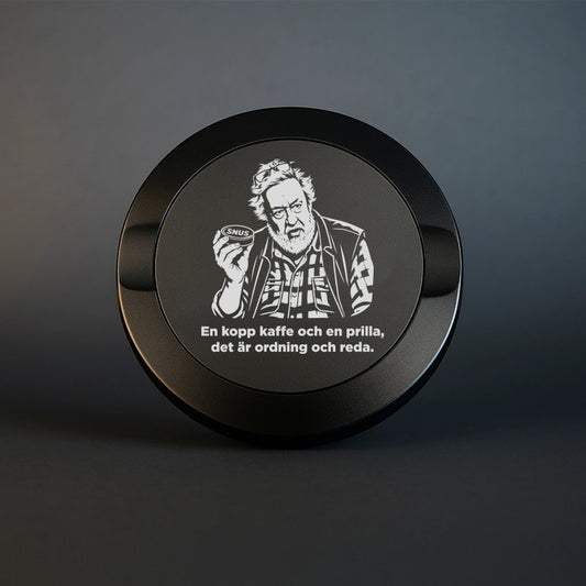 Mattsvart snusdosa i metall med en detaljerad lasergravyr av Leif GW som håller en snusdosa, tillsammans med texten "En kopp kaffe och en prilla, det är ordning och reda".