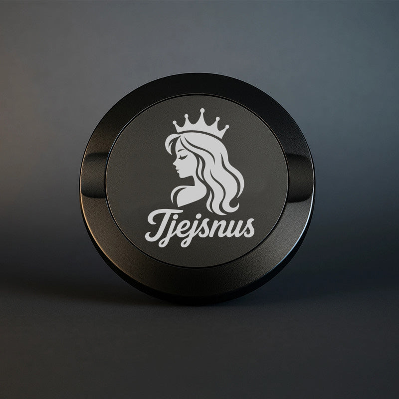 Mattsvart snusdosa i metall med en detaljerad vit gravyr av en kvinna med krona och texten Tjejsnus, fotograferad mot en stilren och mörk bakgrund.